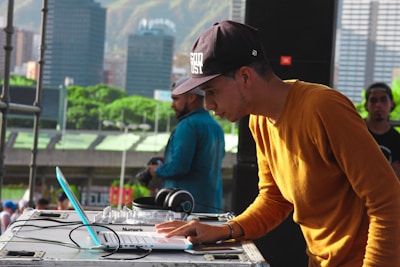 A DJ is focusing on his work at a music event, using a laptop and DJ equipment. He is wearing a mustard-colored long-sleeve shirt and a black cap. In the background, there is a cityscape with tall buildings and green hills.