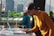 A DJ is focusing on his work at a music event, using a laptop and DJ equipment. He is wearing a mustard-colored long-sleeve shirt and a black cap. In the background, there is a cityscape with tall buildings and green hills.