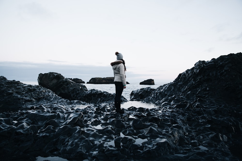 woman in hooded jacket standing on boulder looking at body of water