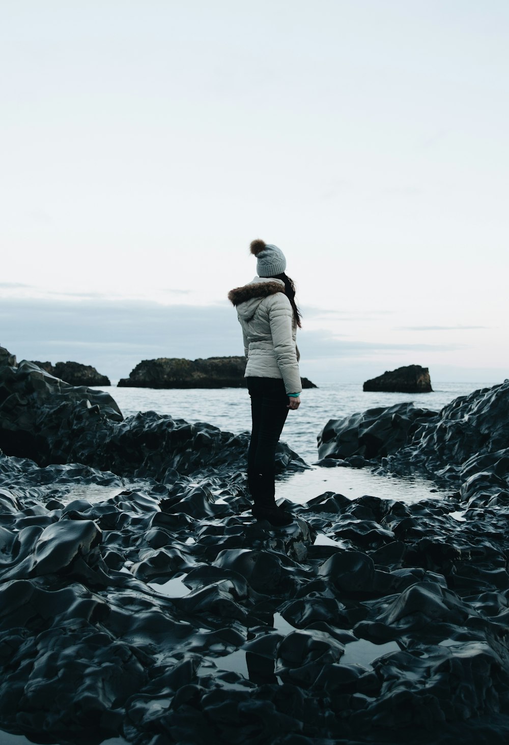 woman in white parka jacket and black pants outfit standing on rocks near body of water under white sky at daytime