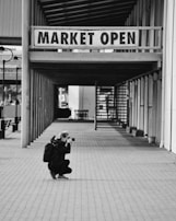 A person is crouching on a paved path, holding a camera up to their face, focused on capturing a photograph. They have a backpack on and are positioned under a sign that reads 'MARKET OPEN' which hangs in a corridor lined with vertical and horizontal beams. The setting has a calm, almost deserted market vibe with structural elements.