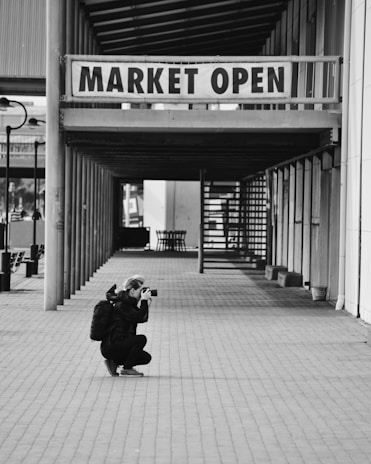 A person is crouching on a paved path, holding a camera up to their face, focused on capturing a photograph. They have a backpack on and are positioned under a sign that reads 'MARKET OPEN' which hangs in a corridor lined with vertical and horizontal beams. The setting has a calm, almost deserted market vibe with structural elements.