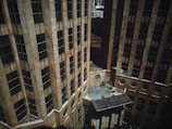 Aerial view of two tall, modern office buildings with beige exteriors and numerous windows. The structures create an urban canyon with a small courtyard between them. Sunlight casts shadows across the facades, adding depth to the scene.