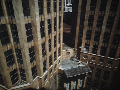 Aerial view of two tall, modern office buildings with beige exteriors and numerous windows. The structures create an urban canyon with a small courtyard between them. Sunlight casts shadows across the facades, adding depth to the scene.