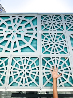 A decorative metal panel with intricate geometric patterns is mounted on a building facade. The panel has a star-like lattice design with teal perforated backing. A hand with raised fingers reaches upwards from the bottom right corner.