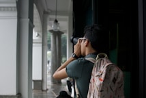 A person is standing under a covered walkway, holding a camera up to take a photograph. The person is wearing a backpack with colorful patterns and a watch on their wrist. The setting appears to be an architectural space with white columns and decorative elements.