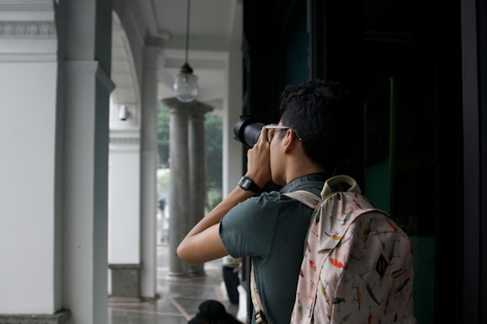 A person is standing under a covered walkway, holding a camera up to take a photograph. The person is wearing a backpack with colorful patterns and a watch on their wrist. The setting appears to be an architectural space with white columns and decorative elements.