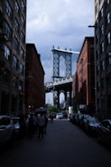 A street view framed by two tall brick buildings with the Manhattan Bridge in the background. Cars line the edges of the cobblestone street, and people are walking or standing on the sidewalks. The sky appears cloudy, contrasting with the industrial structure of the bridge.