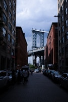 A street view framed by two tall brick buildings with the Manhattan Bridge in the background. Cars line the edges of the cobblestone street, and people are walking or standing on the sidewalks. The sky appears cloudy, contrasting with the industrial structure of the bridge.
