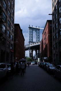 A street view framed by two tall brick buildings with the Manhattan Bridge in the background. Cars line the edges of the cobblestone street, and people are walking or standing on the sidewalks. The sky appears cloudy, contrasting with the industrial structure of the bridge.