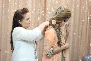 A woman is assisting another woman with her attire. The second woman is wearing an embellished traditional outfit with intricate embroidery and a decorative headpiece adorned with flowers. The background features a shimmering, sequined curtain, adding a glamorous touch to the scene.