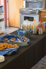 A table is set up with a variety of doughnuts on trays lined with blue napkins, along with serving tongs. There are multiple pitchers with different beverages, including what seems to be orange juice and chocolate milk, with cups arranged nearby. The setting appears to be a kitchen or cafeteria with an oven and other kitchen furniture visible in the background.
