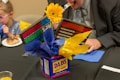 A centerpiece crafted from a Dad's Root Beer container is filled with bright blue and yellow tissue paper, topped with a sunflower. Plaques with humorous messages about house rules and dating are inserted into the centerpiece. A child and an adult are sitting at a table behind it; the adult is taking a bite of food.