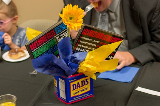 A centerpiece crafted from a Dad's Root Beer container is filled with bright blue and yellow tissue paper, topped with a sunflower. Plaques with humorous messages about house rules and dating are inserted into the centerpiece. A child and an adult are sitting at a table behind it; the adult is taking a bite of food.