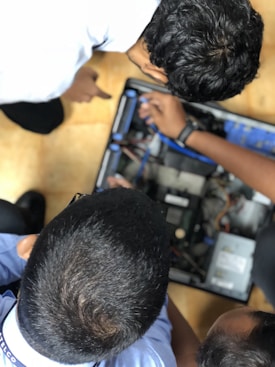 Three individuals are gathered around an open computer case, seemingly engaged in inspecting or repairing the internal components. Their focus is directed towards the intricate circuitry and hardware within. One person's hand is seen pointing towards a specific part, possibly indicating an area of interest or troubleshooting.