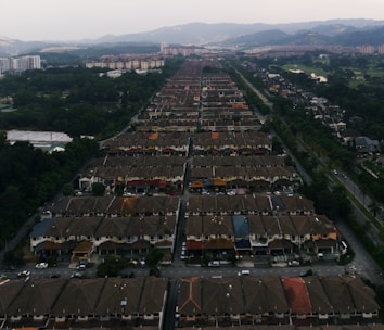 Aerial view of a large residential area featuring rows of similar houses with brown roofs and a range of colored awnings. The houses are lined along parallel streets, surrounded by greenery, with hills and more urban structures in the distance.