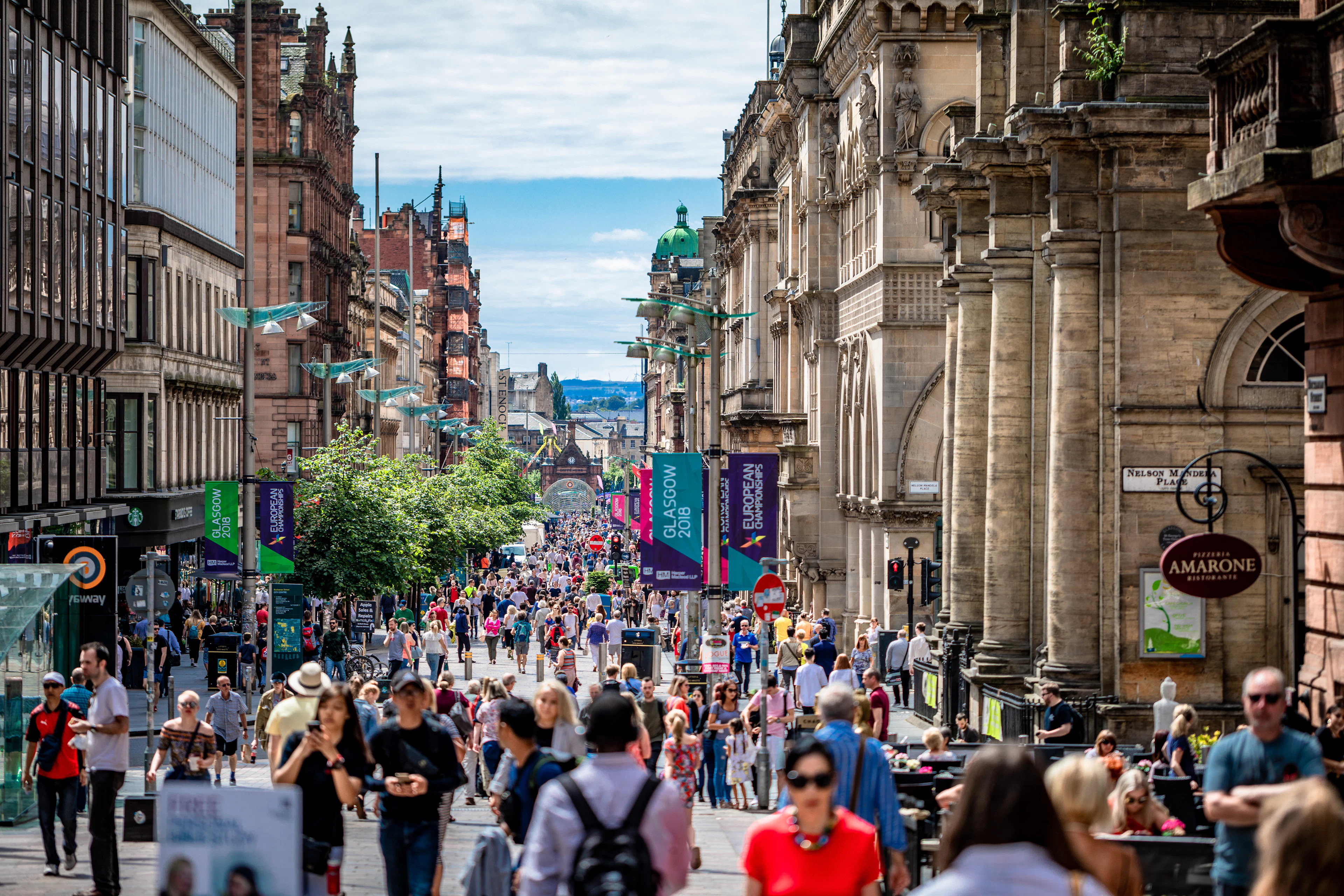 Crowds strolling along a bustling pedestrian shopping street in Glasgow city centre, lined with grand Victorian buildings.