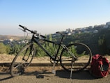 A mountain bike is parked on the side of a road overlooking a lush valley with hills and a scattered urban area in the background. The bicycle stands near a vibrant red backpack, suggesting a pause during a journey. Tall buildings can be seen on the distant hills under a clear, blue sky.