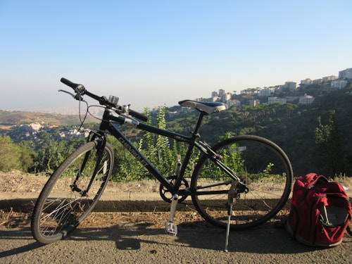 A mountain bike is parked on the side of a road overlooking a lush valley with hills and a scattered urban area in the background. The bicycle stands near a vibrant red backpack, suggesting a pause during a journey. Tall buildings can be seen on the distant hills under a clear, blue sky.