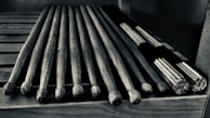 A set of wooden drumsticks are neatly arranged on a slotted wooden surface. There are traditional sticks alongside a bundle of thinner sticks tied together, often used for softer percussion sounds.