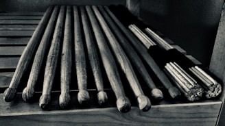 A set of wooden drumsticks are neatly arranged on a slotted wooden surface. There are traditional sticks alongside a bundle of thinner sticks tied together, often used for softer percussion sounds.