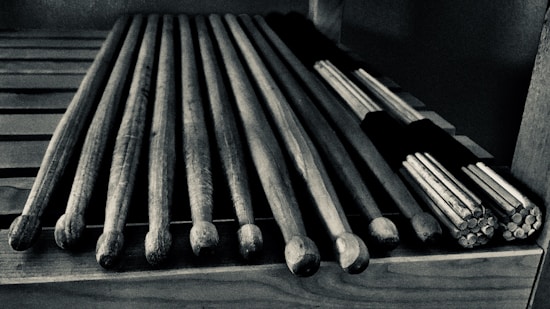 A set of wooden drumsticks are neatly arranged on a slotted wooden surface. There are traditional sticks alongside a bundle of thinner sticks tied together, often used for softer percussion sounds.