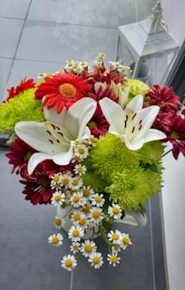 A vibrant bouquet of flowers featuring white lilies, red and maroon chrysanthemums, green pompoms, and small white daisies with yellow centers. The arrangement is in a white vase, set on a tiled floor with a decorative lantern nearby.