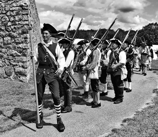 A group of individuals dressed in historical military attire from the 18th century are marching along a pathway beside a stone wall. Each person is wearing a tricorne hat and carrying muskets. The scene appears to be a reenactment of a historical event, possibly a battle or military drill. The setting is outdoors under a partly cloudy sky, with grass and trees in the background.