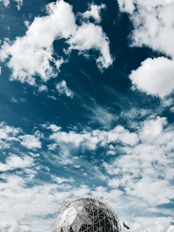 A geodesic dome structure is positioned against a vibrant sky filled with scattered white clouds. The architectural design features triangular patterns with a metallic sheen, creating a striking contrast with the deep blue and white of the sky.