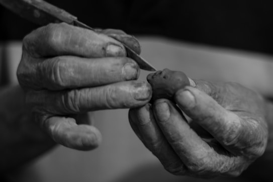 A pair of weathered hands are carefully sculpting a small piece of clay using a tool. The fingers are slightly dirty, indicating involvement in a hands-on craft activity.