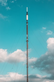 A tall, slender broadcast tower stretches into the sky, supported by numerous cables. The sky is mostly clear with a few fluffy clouds scattered around.