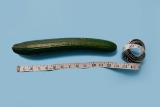 A long, green cucumber lies flat on a light blue background beside a white measuring tape. The measuring tape is extended alongside the cucumber to show length, with part of it coiled in a roll.