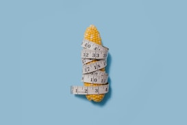 A yellow corn cob is wrapped around with a white measuring tape. The background is a solid light blue color, providing a stark contrast to the corn. The tape measure is coiled in several loops, displaying numbers clearly visible on its surface.