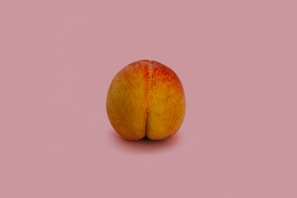 A single peach is placed against a plain pink background. The fruit is prominently displayed in the center, with visible textures and colors that range from yellow to red.