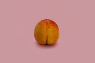 A single peach is placed against a plain pink background. The fruit is prominently displayed in the center, with visible textures and colors that range from yellow to red.
