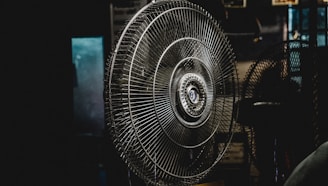 A large metal fan placed indoors, with its intricate wire grille and central hub prominently featured. The setting is dimly lit, creating a moody atmosphere with shadows surrounding the fan.