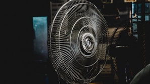 A large metal fan placed indoors, with its intricate wire grille and central hub prominently featured. The setting is dimly lit, creating a moody atmosphere with shadows surrounding the fan.