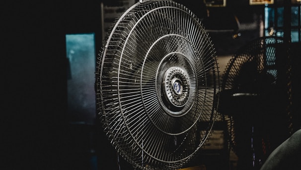 A large metal fan placed indoors, with its intricate wire grille and central hub prominently featured. The setting is dimly lit, creating a moody atmosphere with shadows surrounding the fan.