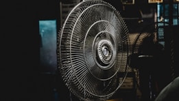 A large metal fan placed indoors, with its intricate wire grille and central hub prominently featured. The setting is dimly lit, creating a moody atmosphere with shadows surrounding the fan.