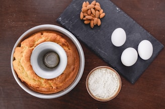 A bundt cake in a baking pan is placed alongside a black slate board holding a handful of almonds and three eggs. A small bowl of flour is also on the table, all set on a dark wooden surface.