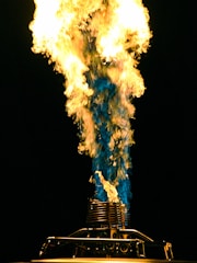 A large, brightly colored flame shoots upward, displaying a mix of yellow, orange, and blue hues. The flame originates from a metal contraption, likely a burner or torch, with intricate details visible at the base.