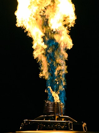 A large, brightly colored flame shoots upward, displaying a mix of yellow, orange, and blue hues. The flame originates from a metal contraption, likely a burner or torch, with intricate details visible at the base.