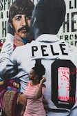 A mural featuring a person in a patterned jacket alongside another wearing a shirt with the name 'Pelé' on it. In front of the mural, there is a woman in a red and white striped dress, looking to the side.