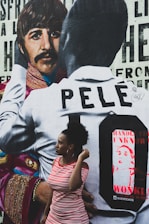 A mural featuring a person in a patterned jacket alongside another wearing a shirt with the name 'Pelé' on it. In front of the mural, there is a woman in a red and white striped dress, looking to the side.