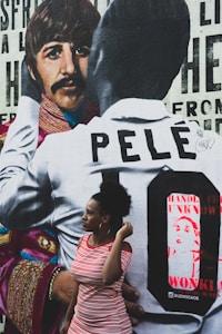 A mural featuring a person in a patterned jacket alongside another wearing a shirt with the name 'Pelé' on it. In front of the mural, there is a woman in a red and white striped dress, looking to the side.