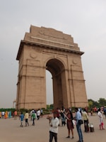 A large stone archway stands prominently in an open area with several people walking and standing around. The archway is ornate and has 'India' inscribed at the top. People are dressed in various styles, with some taking photos and others walking by.