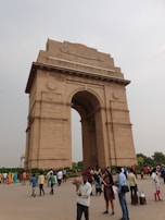 A large stone archway stands prominently in an open area with several people walking and standing around. The archway is ornate and has 'India' inscribed at the top. People are dressed in various styles, with some taking photos and others walking by.