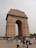 A large stone archway stands prominently in an open area with several people walking and standing around. The archway is ornate and has 'India' inscribed at the top. People are dressed in various styles, with some taking photos and others walking by.