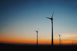 Silhouettes of wind turbines stand against a vibrant sunset sky, transitioning from a deep orange near the horizon to a clear blue as it rises.