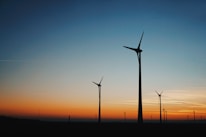 Silhouettes of wind turbines stand against a vibrant sunset sky, transitioning from a deep orange near the horizon to a clear blue as it rises.